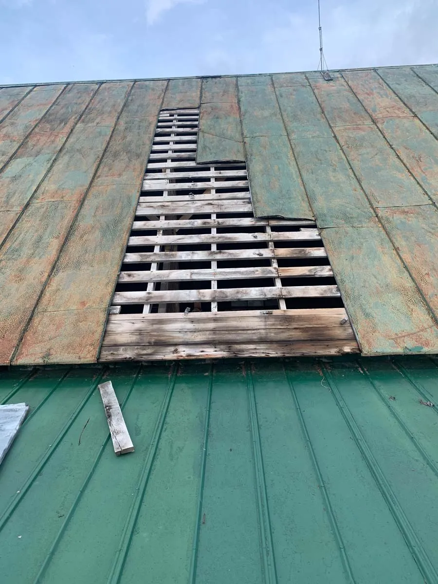 Storm-damaged metal barn roof with missing panels exposing the wood roof boards beneath